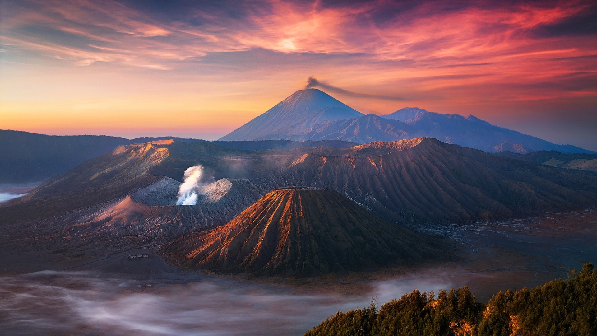 Matahari terbit di kaldera Tengger, Semeru mengepul di belakang Bromo dan Batok, awan memenuhi dasar kaldera.